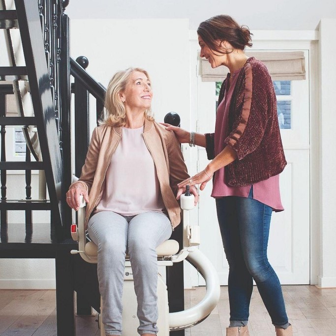 Lady riding Curved Stairlift with another lady assisting.